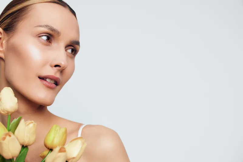 Young woman with clear skin holding yellow tulips close to her shoulder, soft natural light, gentle expression, minimal makeup, plain light background.