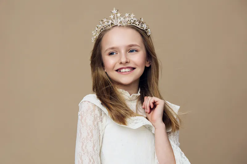 Portrait of a young girl wearing a delicate crown, smiling joyfully The soft background enhances her playful demeanor and elegant white dress, perfect for royalty themes