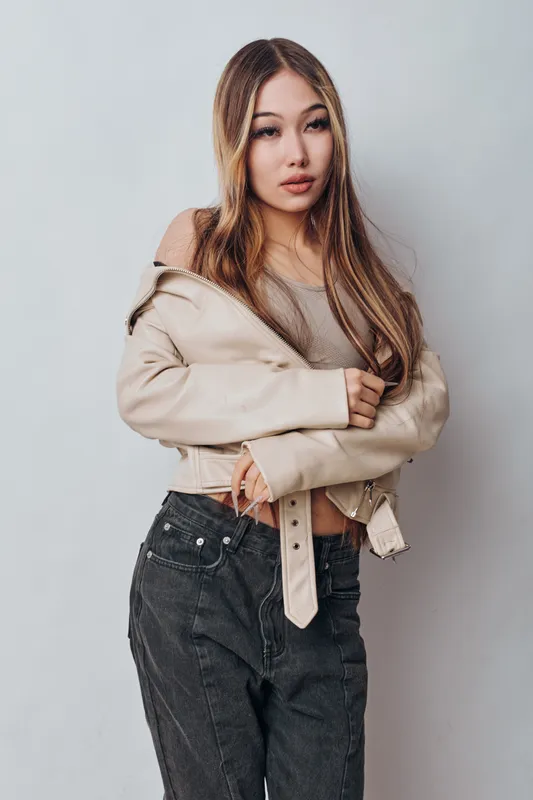Young woman with long hair posing in beige jacket and black jeans, looking confident and stylish against plain background with casual fashion outfit.