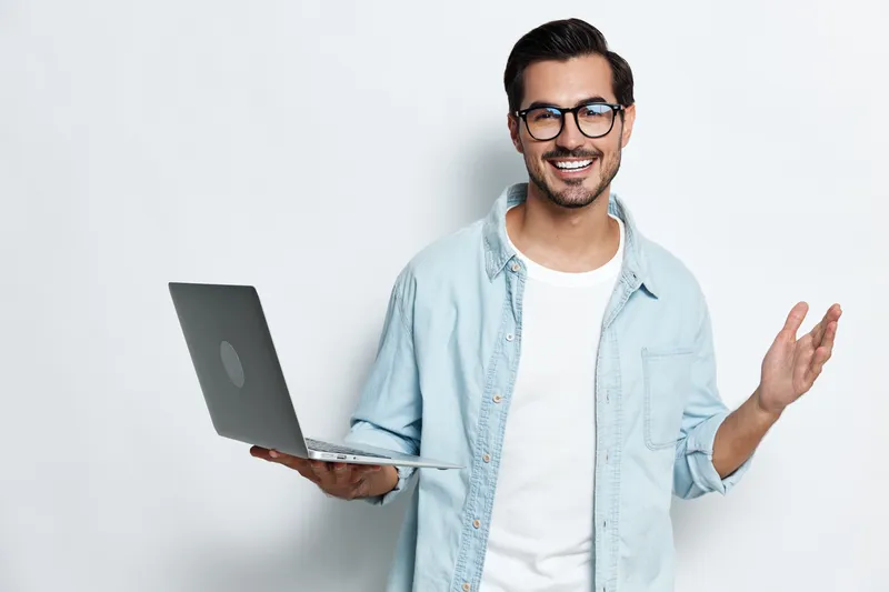 Smiling young man wearing glasses and a denim shirt holds a laptop while gesturing casually, looking at the camera with a cheerful expression against a light backdrop.