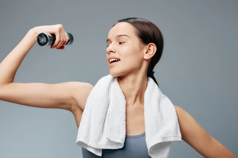 Female fitness enthusiast lifting a dumbbell with a towel around the neck, in a gym setting, showcasing strength, focus, and athletic movement during a workout.