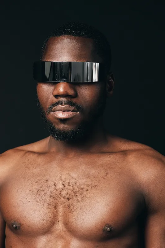African man with dark skin and beard wearing futuristic reflective visor in creative conceptual visual style on black background. Shirtless male portrait with modern tech accessory and confident