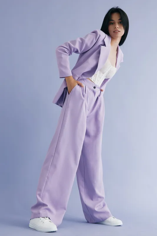 stylish woman wearing lilac suit and white sneakers poses confidently against pastel background, showcasing modern high-waisted wide-leg pants and casual fashion style