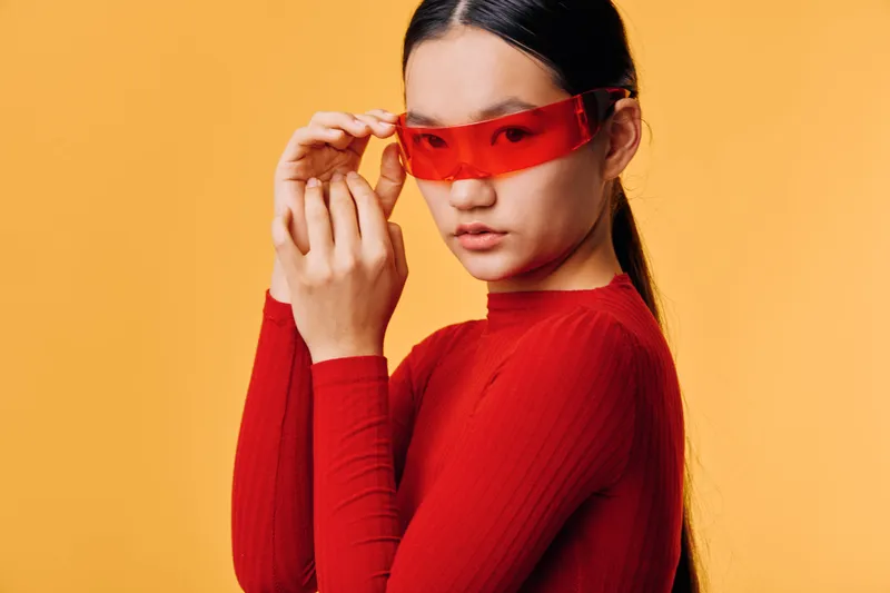 Young woman in red long sleeve shirt adjusting red transparent sunglasses standing against orange background for fashion and style portrait.