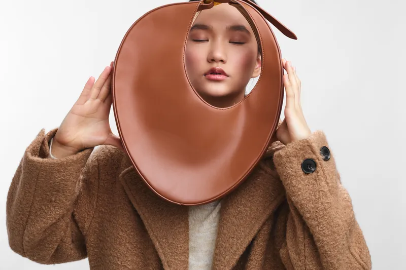 asian woman wearing a brown wool coat holds a circular leather neck accessory near her face, creating a bold fashion statement and a sophisticated contemporary look.