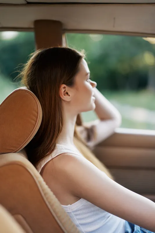 Young woman relaxing in a car with her hand on her head, enjoying a sunny day outdoors, warm tones, casual clothing.