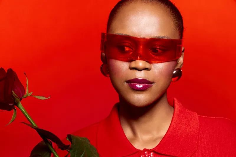 Portrait of a dark skinned African American woman wearing red sunglasses and red clothing holding a red rose on a vibrant red background. Concept of fashion, beauty, and elegance.