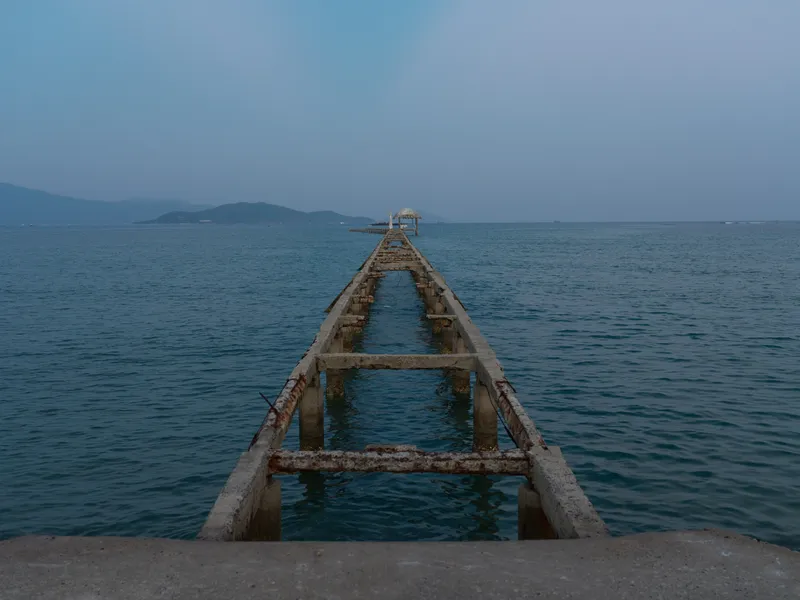 rusty pier extending into calm blue sea during twilight with distant mountains on horizon