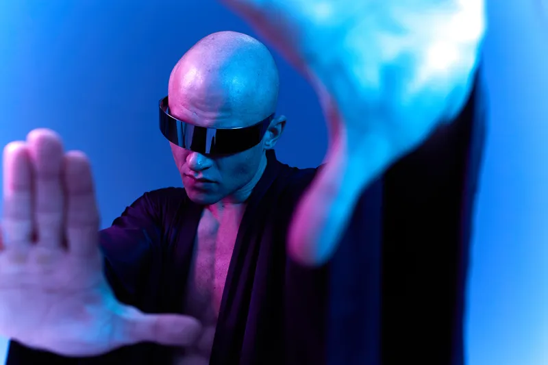 Stylish bald man in a futuristic outfit poses dramatically with blue lighting against a vibrant background, showcasing individual expression and modern aesthetics.