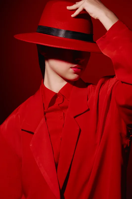 Young woman in a red oversized coat and matching hat partially covers her face. Fashion concept with bold color contrast and dramatic shadows for beauty editorial.