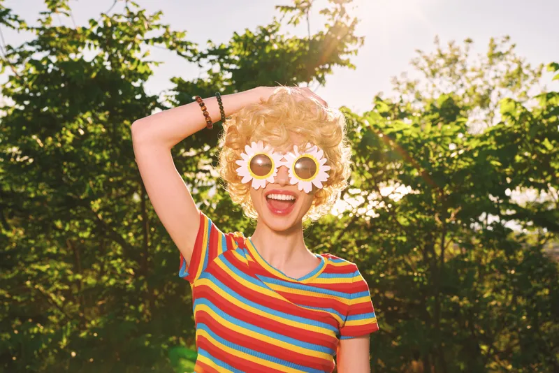 A joyful woman in a colorful striped shirt poses outdoors in bright sun, wearing playful flower sunglasses. A retro film color concept highlights carefree style and whimsy.