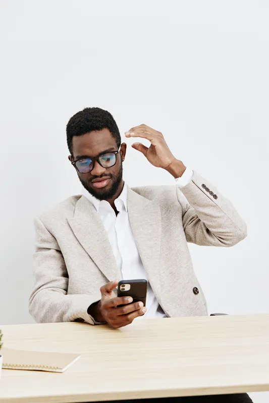 Professional young African-American man in stylish beige blazer and glasses using mobile phone while sitting at a desk against plain white background, studio portrait. Technology work concept