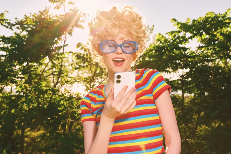 Vibrant woman outdoors wearing a striped colorful shirt and retro sunglasses, capturing a candid lifestyle moment with warm sun, bright colors, and a playful, carefree mood.