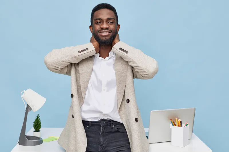 Smiling man wearing light gray sweater and white shirt looking confident and relaxed at home workspace with desktop lamp plant and laptop isolated on light blue background modern lifestyle concept