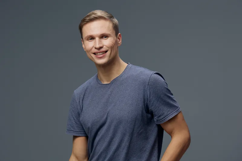 Smiling young man wearing casual blue t-shirt against plain gray background, looking at camera, happy expression, pose indoors, natural lighting, portrait shot, youthful appearance, relaxed mood.