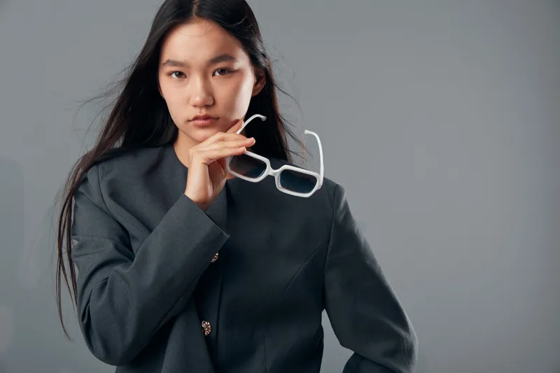 Portrait of confident young woman in dark blazer holding white eyeglasses with long hair against neutral gray background. Serious expression, studio lighting and minimalistic fashion style.