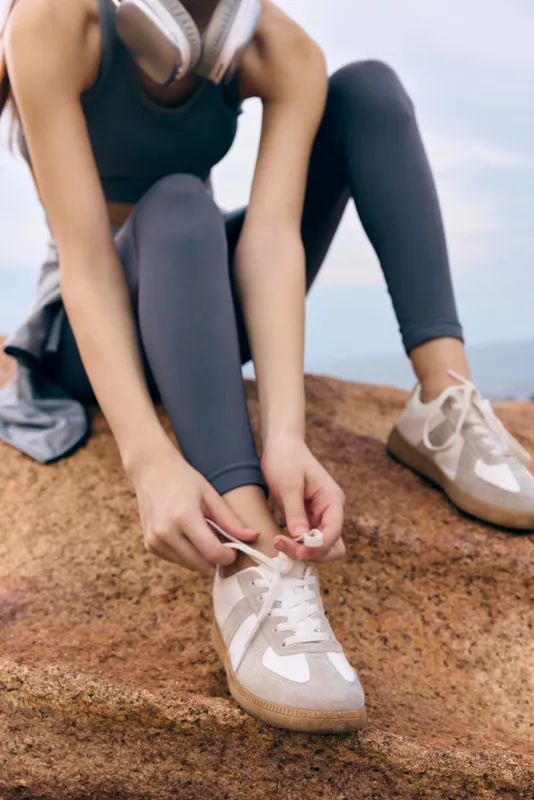 Young woman tying shoelaces on athletic shoes, wearing fitness apparel, with headphones, outdoors against a natural backdrop, focused and determined