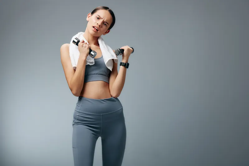 Woman in athletic wear holds dumbbells with a towel over her shoulders, posing in a studio. A strong fitness portrait highlighting strength, balance and focus during training.