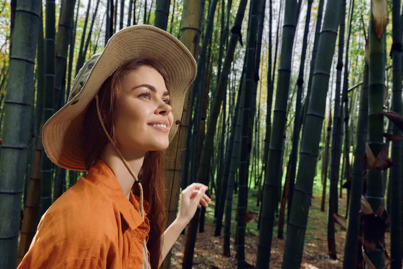 Woman in hat smiling among tall bamboo forest, portrait side view with sunlight filtering through green foliage. Nature travel moment, orange shirt, serene outdoors explorer in grove.
