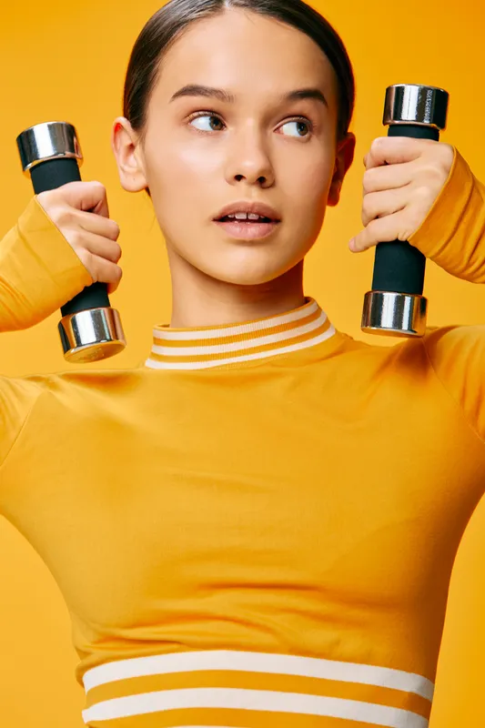 Fitness minded woman in a yellow athletic top lifts dumbbells against a bright orange backdrop, showcasing strength, focus and proper form during a dynamic workout session.