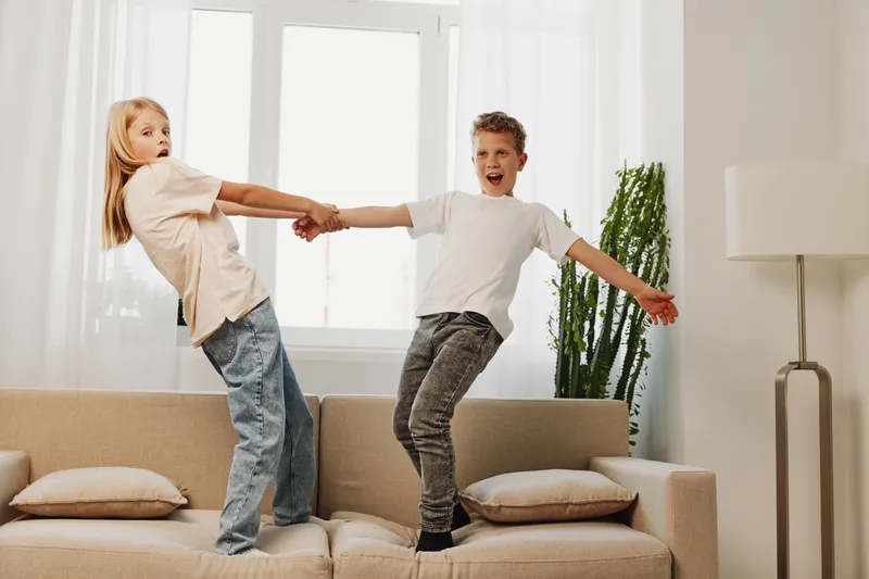 Two playful kids holding hands and balancing on a beige couch in a bright living room with natural light and indoor plants. Concept of childhood fun, friendship, and home life.