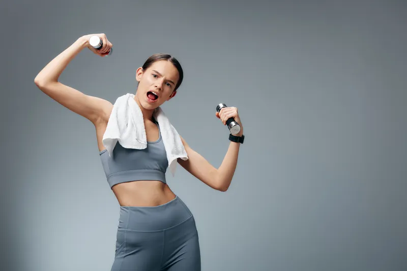 Fitness woman in athletic wear lifts small dumbbells with a towel over her shoulders, mid workout pose, energetic expression, minimal studio backdrop, dynamic fitness scene.