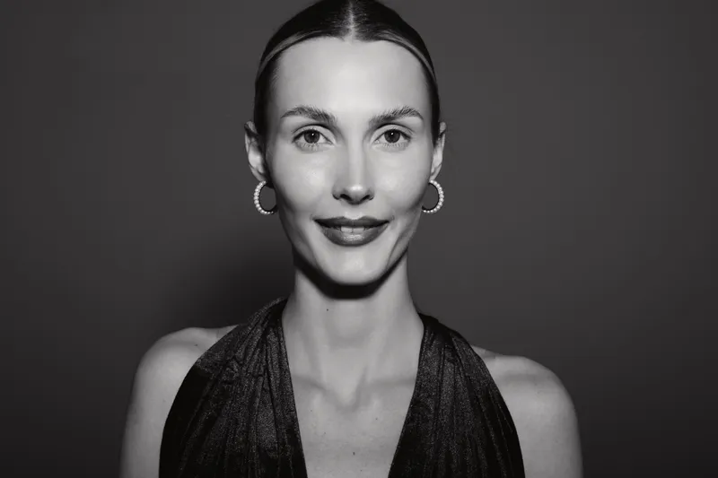 Portrait of a slender white woman with light skin and slicked hair wearing earrings and a dark dress, smiling gently on a dark background.