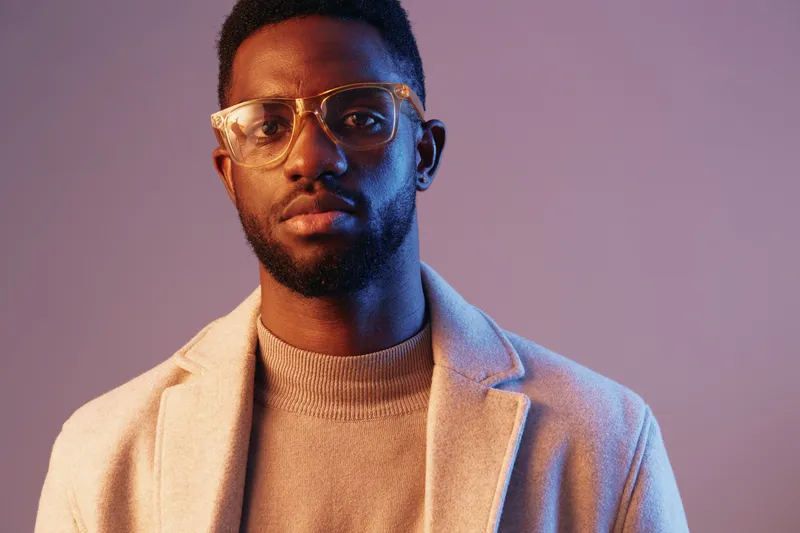 Stylish young man with glasses posing in a soft pastel background, showcasing modern fashion, confidence, and sophistication
