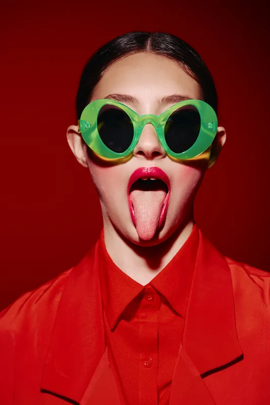 Young woman with large green sunglasses wears bright red clothing and bold lipstick while sticking out her tongue against a solid red background. Concept of beauty-clash and fashion-creativity.