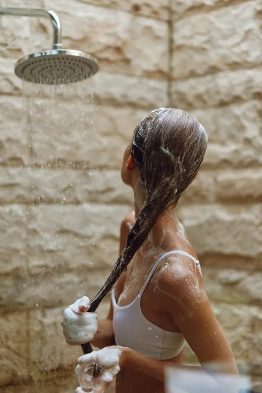 woman washing hair in shower with soap foam, textured stone wall background, relaxed self-care moment, natural light, indoor bathroom scene