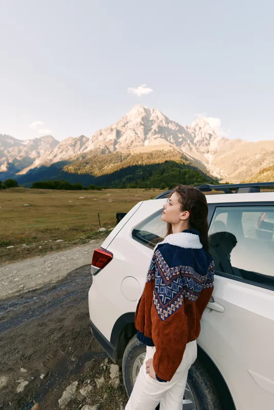Woman leaning on a white car beside a mountain landscape and road, wearing a patterned sweater for travel and outdoors adventure, enjoying nature and scenic autumn light.
