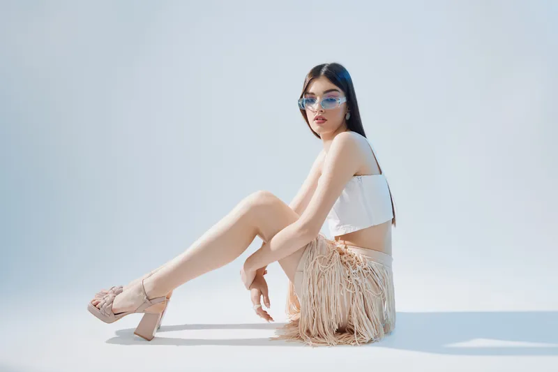 Young woman in stylish sunglasses and fashion outfit poses sitting on white floor. Concept of beauty, fashion, and modern lifestyle with soft natural light.
