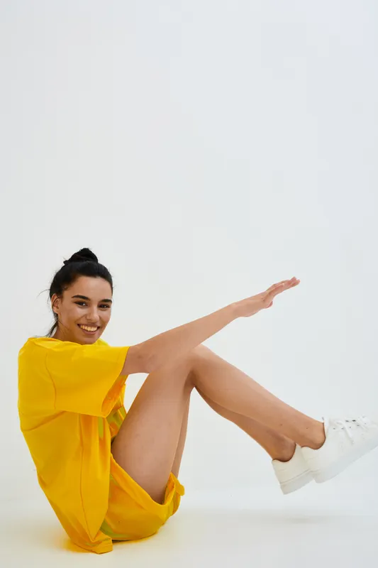 smiling woman in yellow dress sitting on floor with white sneakers, casual lifestyle concept, studio shot with minimal white background