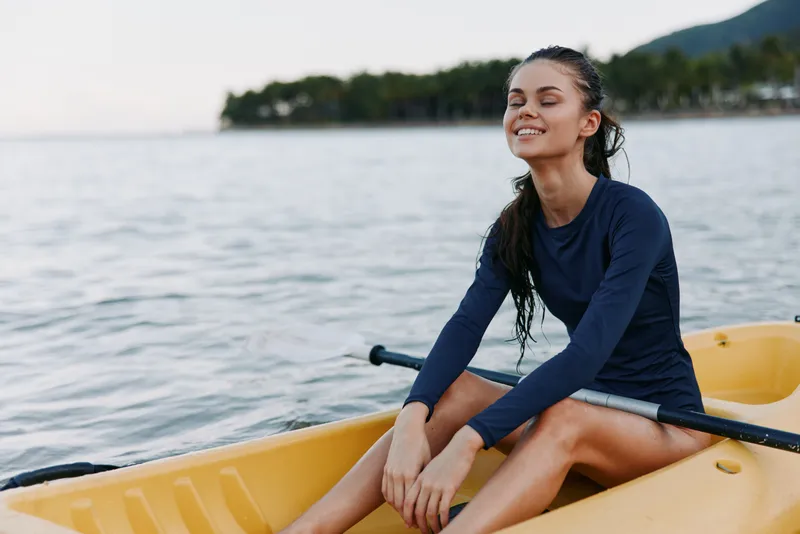 A young woman sits in a bright yellow rowboat on calm water, wearing a navy long-sleeve top and smiling with eyes closed, enjoying a peaceful outdoor moment.