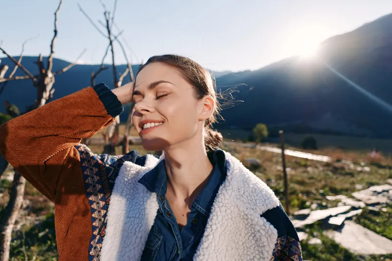 Woman smile outdoors sunlight mountains portrait with closed eyes, young female enjoying countryside in a cozy jacket and sweater during golden hour, peaceful travel lifestyle moment.