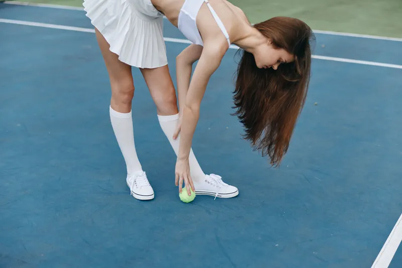 Young female tennis player in white sportswear, bending down to pick up a tennis ball on a blue court, showcasing athleticism and focus