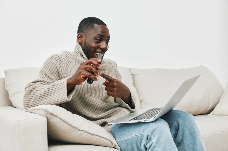 A joyful young Black man, wearing a cozy sweater, enjoys vaping while sitting on a sofa with his laptop, evoking relaxation and modern lifestyle