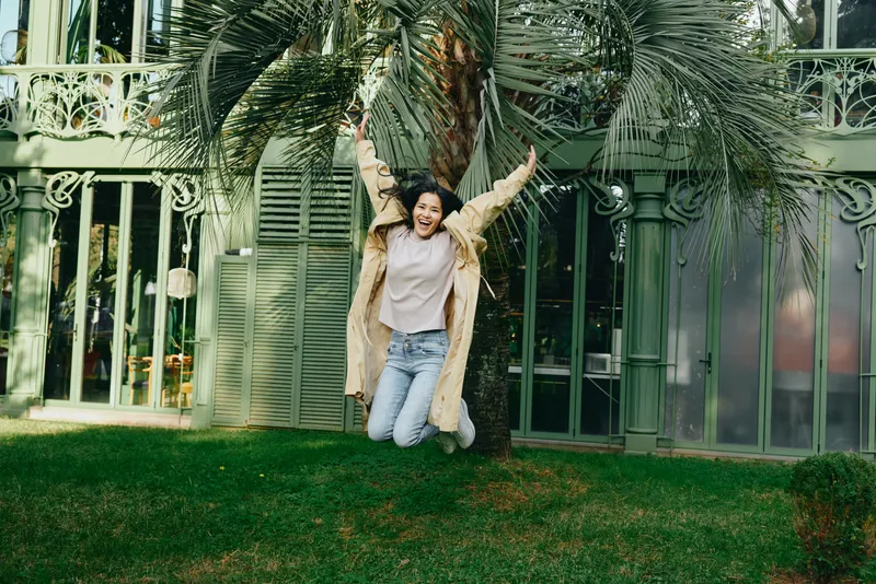 Joyful jump for outdoors scene with a smiling woman in a beige coat, lush garden setting, palm trees, and a decorative fence, capturing carefree happiness and natural movement