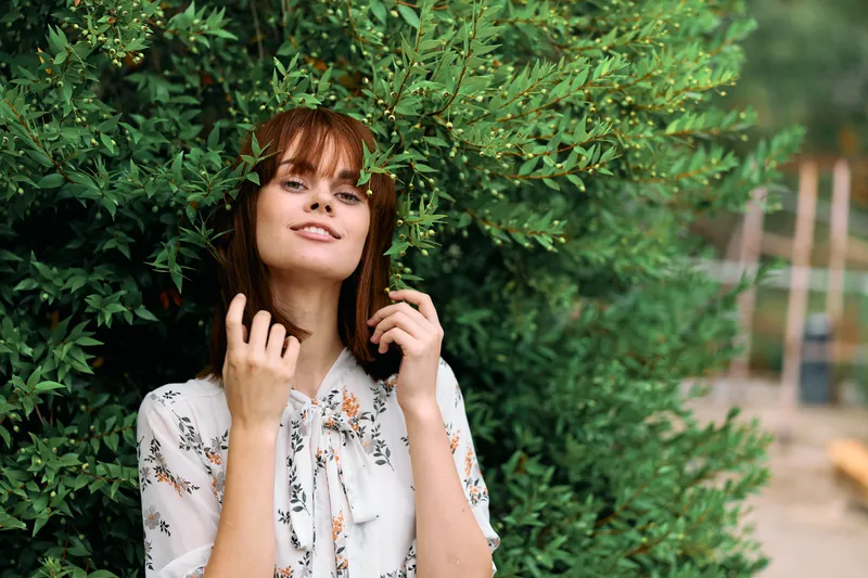 Счастливая женщина на фоне куста, держа руки на голове и улыбаясь