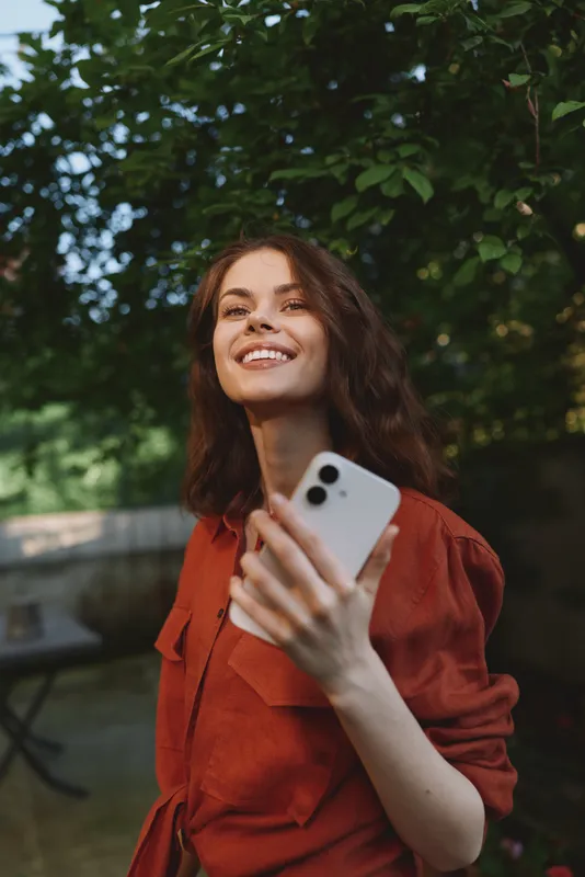 Cheerful woman outdoors, smiling with phone in hand, sunlit moment, casual red outfit, relaxed posture, soft breeze, natural colors, lush greenery background, candid lifestyle portrait