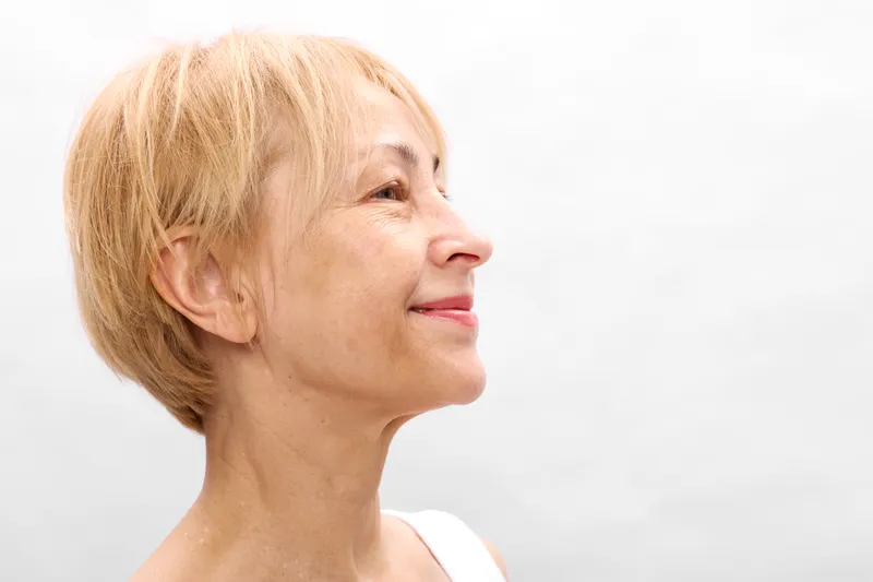 Smiling elderly woman in profile with short blonde hair against a white background. Close-up portrait highlights calm wellness, healthy aging, and gentle self care. Soft studio lighting.