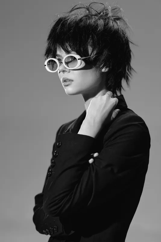 Black and white portrait of a stylish woman wearing white sunglasses with short dark hair and a pensive expression.