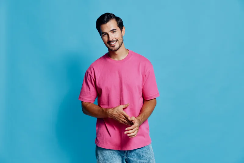 Full body smiling cheerful young man wearing pink t-shirt poses confidently against a solid blue background, showcasing an upbeat lifestyle.
