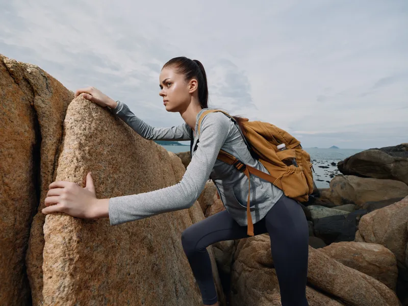 Young woman climbing on rocky coastal terrain, wearing athletic clothing and a backpack, showcasing determination and adventure in a picturesque outdoor setting