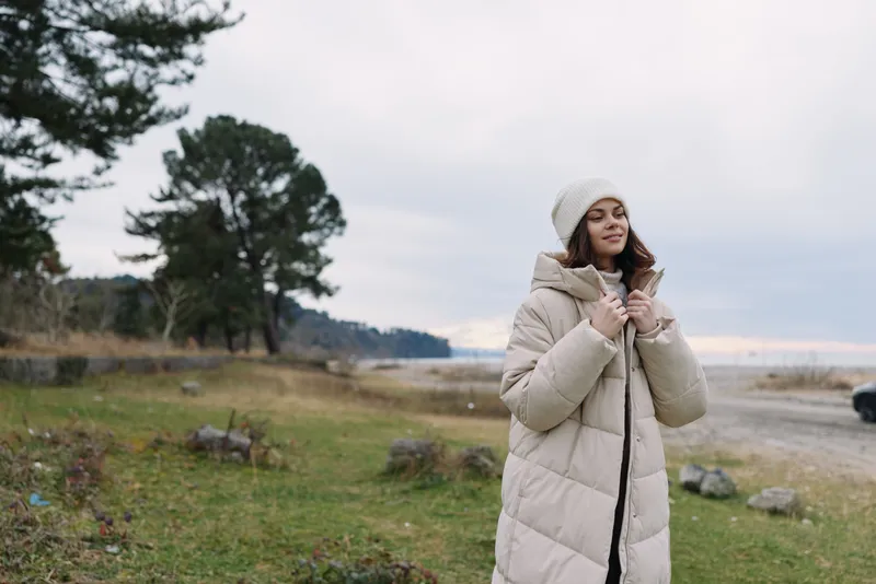 Woman in a plush beige winter coat stands outdoors in a grassy landscape, adjusting her beanie, capturing a calm mood, soft light and distant trees under a cloudy sky.