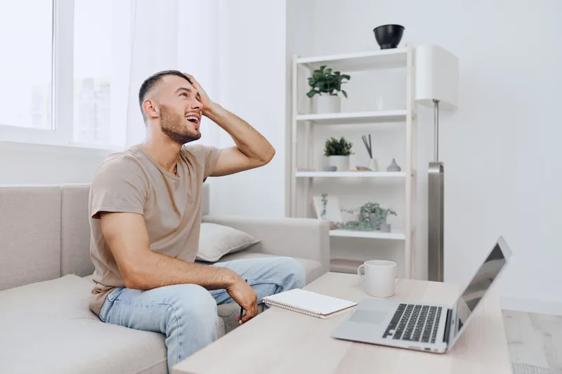 Young man expressing frustration while working from home, dressed casually, surrounded by modern decor, and a calm atmosphere. Ideal for remote work themes.