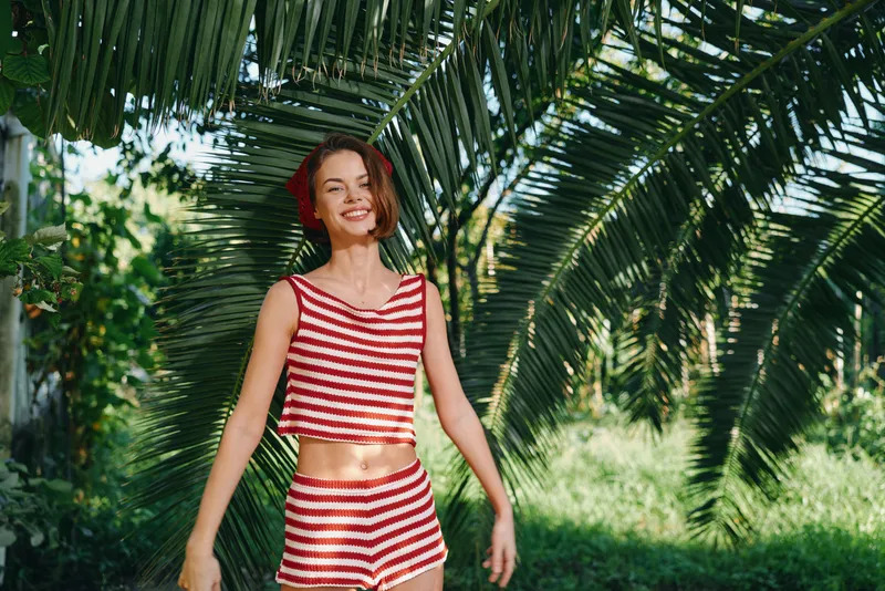 A cheerful young woman stands among tropical palms in a sunny outdoor setting wearing a red and white striped swimsuit top and shorts, smiling and relaxed.