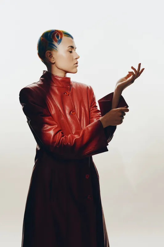 Woman with colorful short hair wearing a red leather coat, striking a thoughtful pose against a plain bright background, showcasing contemporary fashion style.