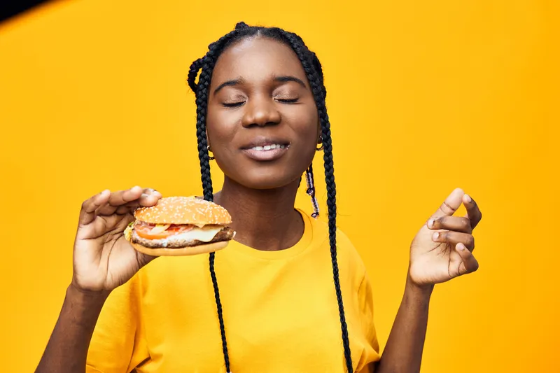 Young Black woman enjoying a delicious burger against a vibrant yellow background, conveying joy and satisfaction in casual summer attire