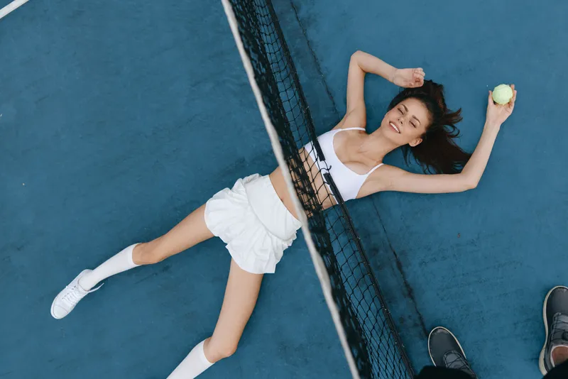 A joyful young woman resting on a tennis court, wearing sporty attire and holding a tennis ball, showcasing a lively and energetic atmosphere